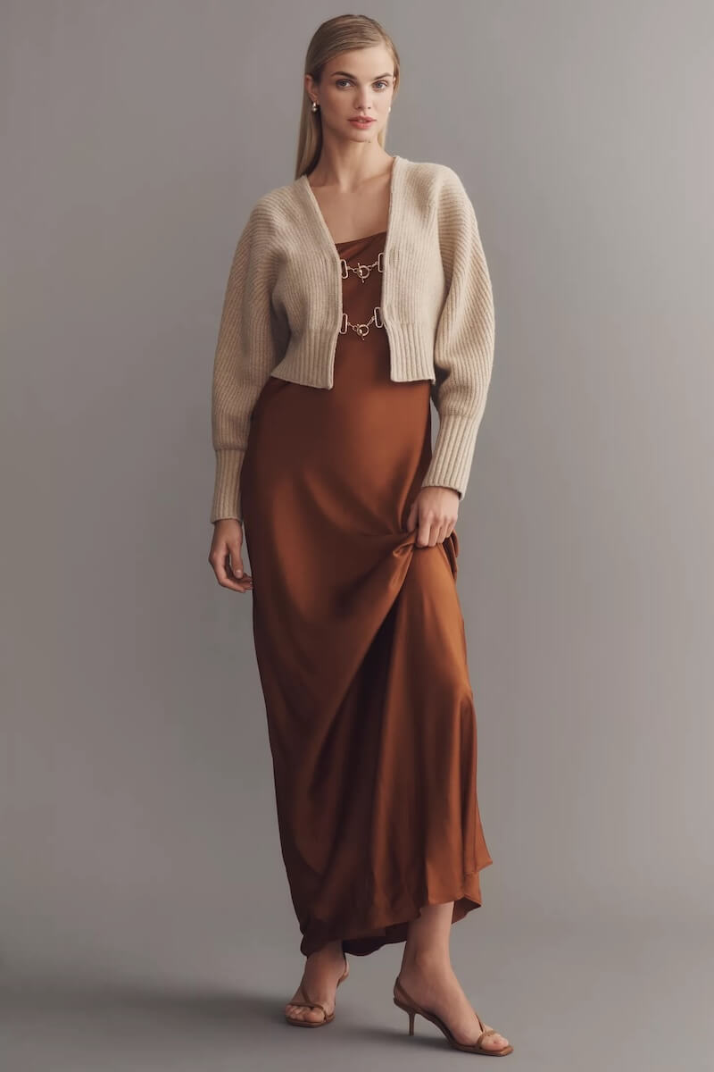 Fall 2025 Fashion: A Stylist's Lookbook of This Season's Trends - 2 A woman stands in front of a plain gray background, wearing a long brown dress, a cropped beige cardigan, and heeled sandals—an effortlessly chic look inspired by fall 2025 fashion.