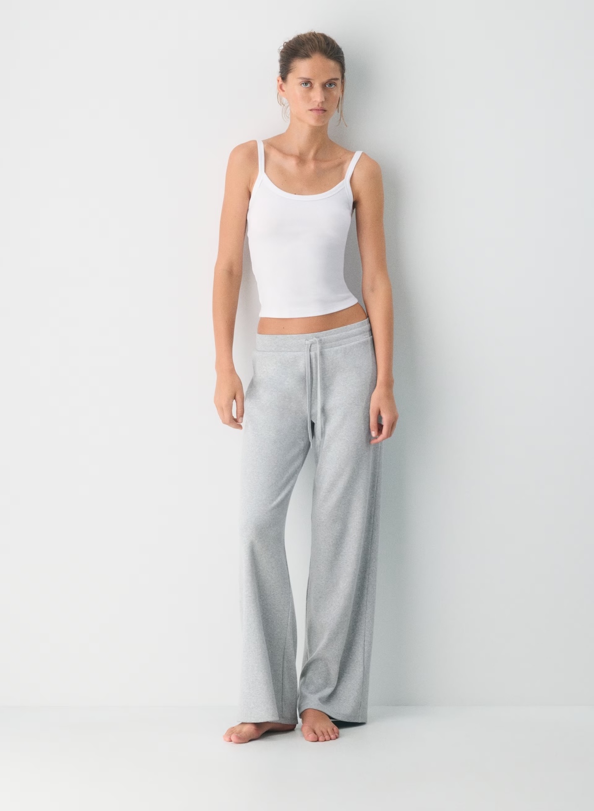 A woman stands barefoot against a plain white background, wearing a white tank top and light gray drawstring lounge pants—an effortless look perfect for the SB Hot List August.