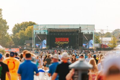 A large crowd gathers in front of the Bourbon & Beyond festival stage during the day, with trees and tents visible in the background—a vibrant scene that captures the spirit of Louisville events in September.