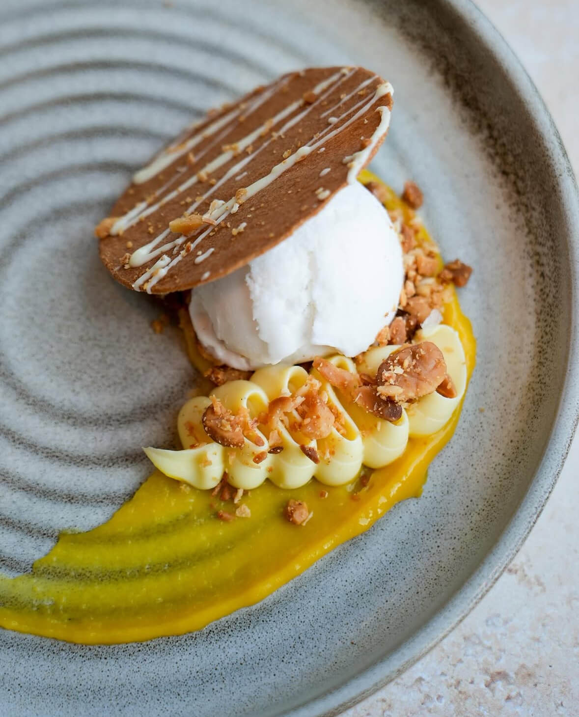 A plated dessert from new menus at Louisville restaurants featuring a scoop of ice cream, a thin crisp cookie, crumbled nuts, and piped yellow cream on a gray spiral-patterned plate.