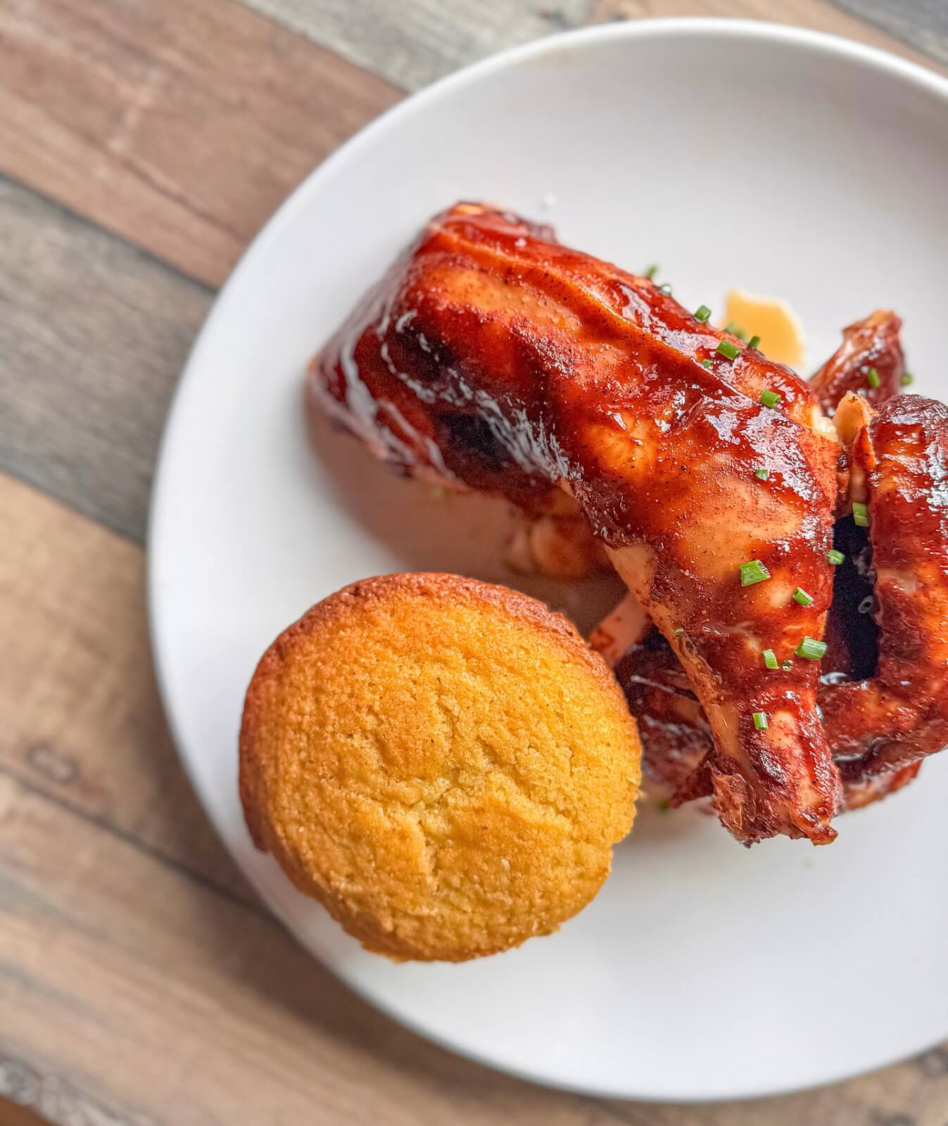 Barbecued chicken with sauce and herbs served on a white plate, accompanied by a round cornbread muffin on a wooden table.