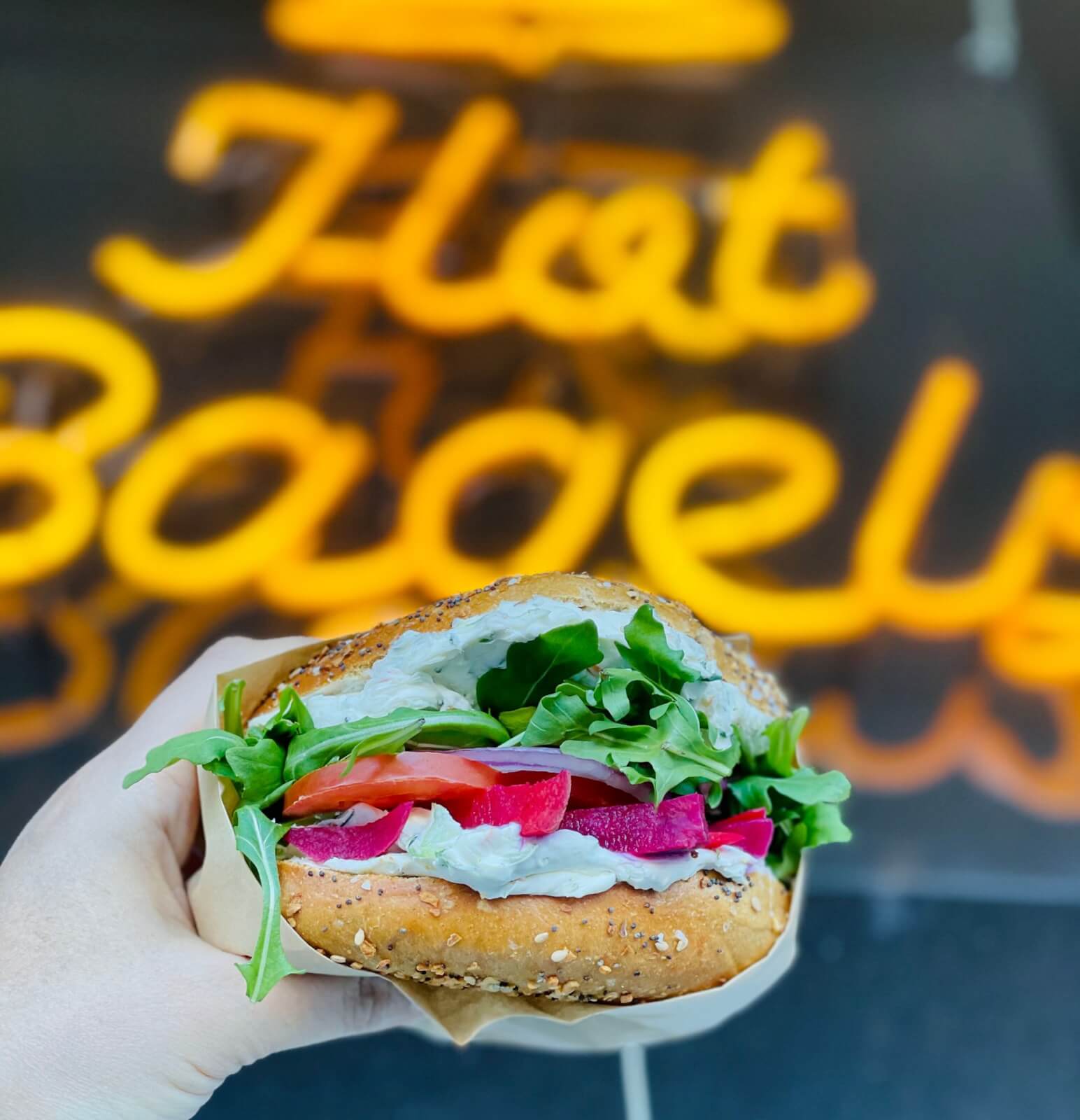 A hand holds a bagel sandwich filled with cream cheese, lettuce, tomato, and pickled onions in front of a blurred neon "Hot Bagels" sign.