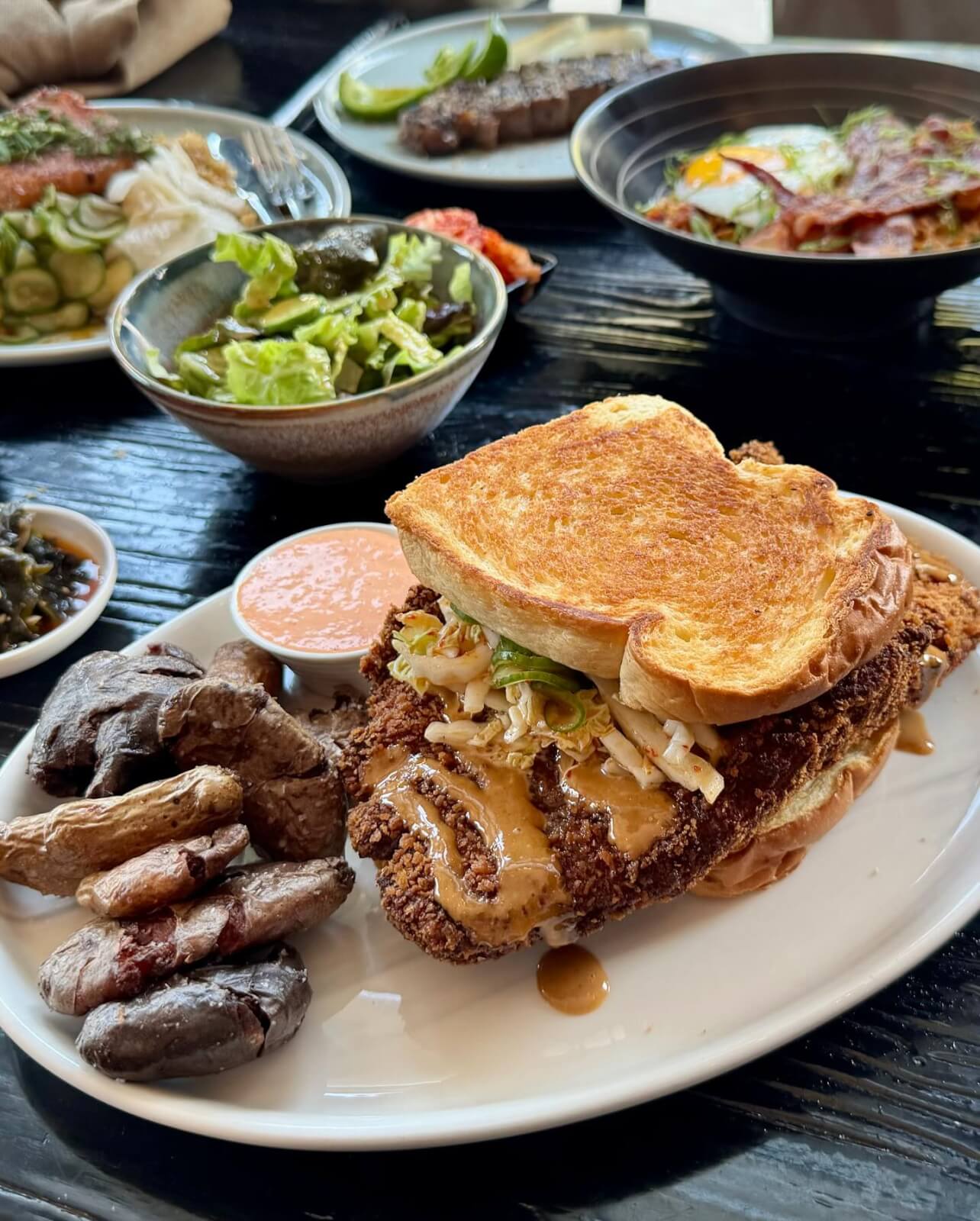 A plate with a fried chicken sandwich on toasted bread, roasted fingerling potatoes, sauce, and a side of lettuce salad—perfect for discovering new menus at Louisville restaurants; other dishes can be seen in the background.