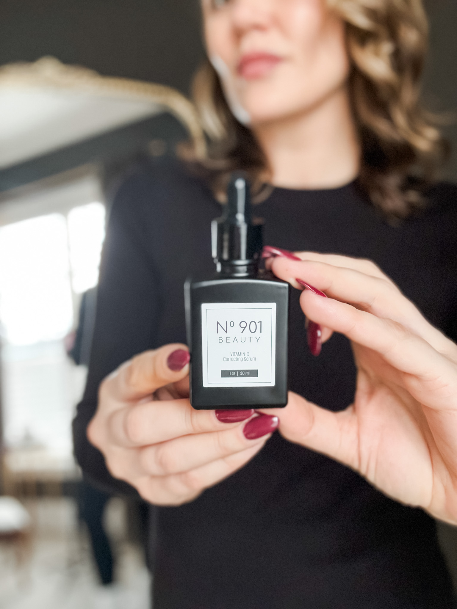 A person with manicured nails holds a black "No 901 Beauty" bottle with a dropper cap, while standing indoors.