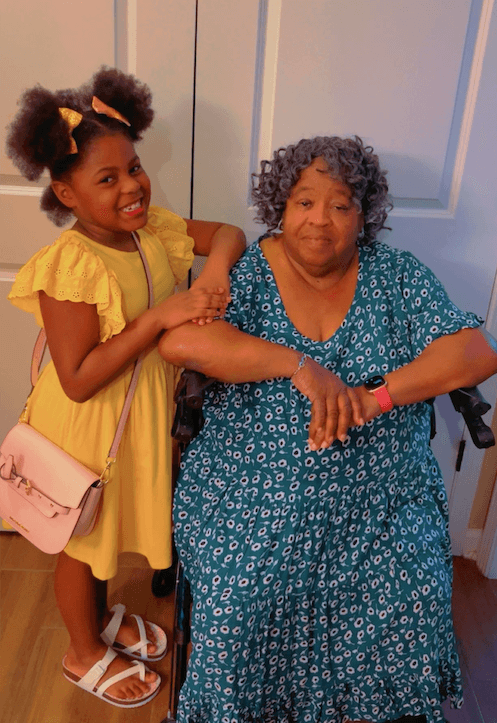 A young girl in a yellow dress stands and smiles beside an older woman with gray curly hair seated in a wheelchair, both posing indoors—capturing a heartwarming moment.