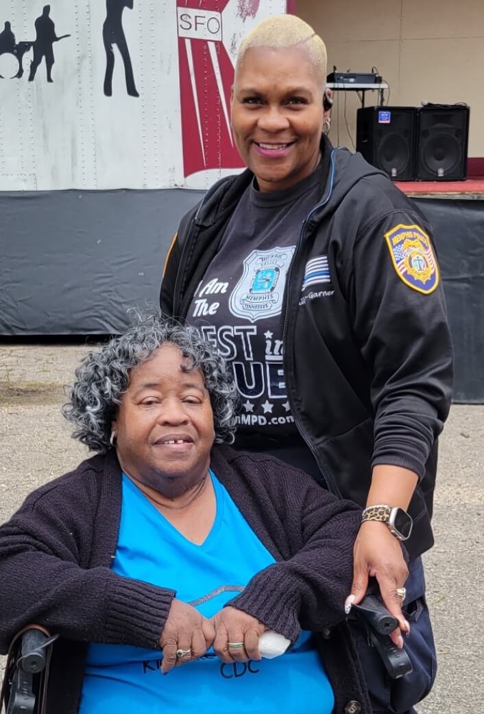 Two women pose outdoors; one is seated in a wheelchair wearing a blue shirt, while the other stands behind her smiling in a black jacket with a police patch.