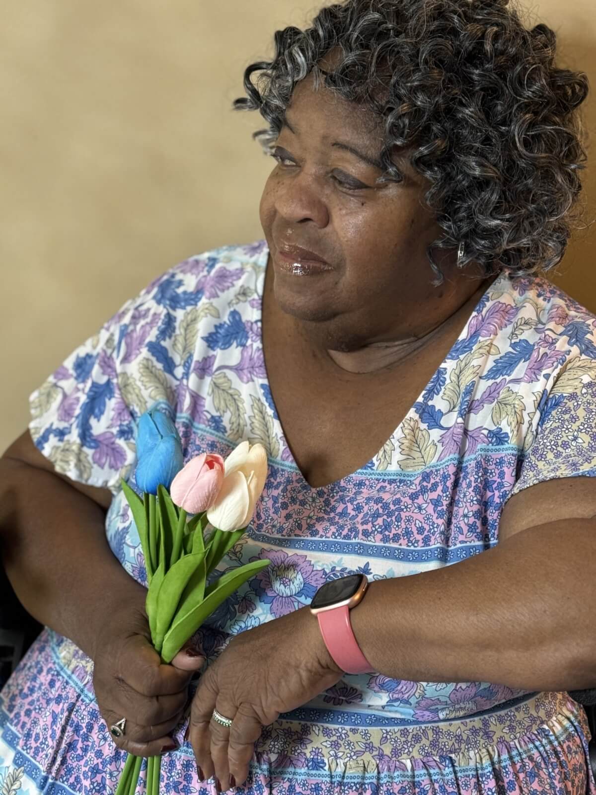 An older woman with curly gray hair holds a small bouquet of tulips and wears a patterned dress and a pink smartwatch.