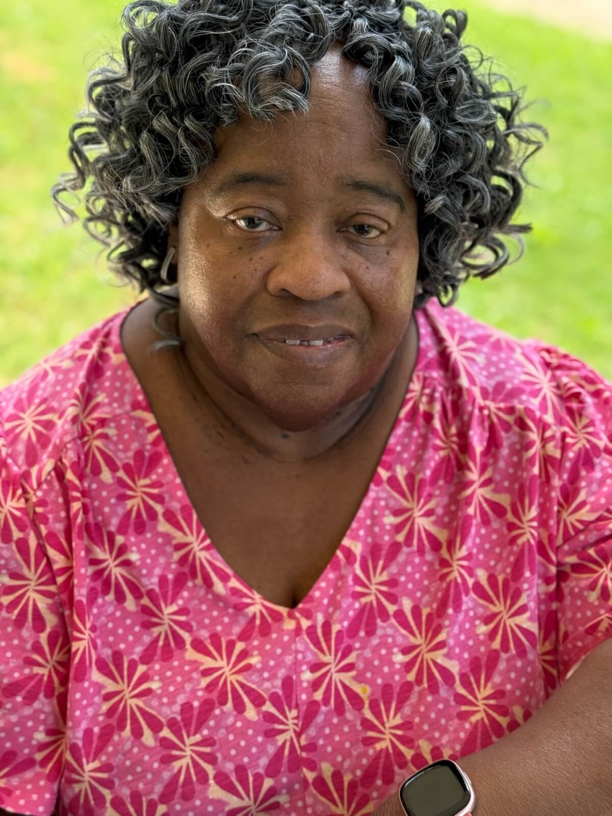 A woman with curly gray hair, wearing a pink patterned blouse and a smartwatch, sits outdoors on a grassy background.