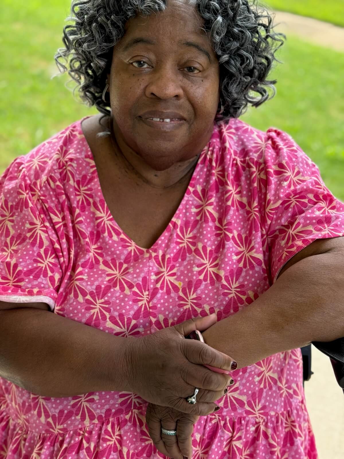 An older woman with curly gray hair sits outdoors, wearing a pink patterned dress and resting her arms on a walker. 