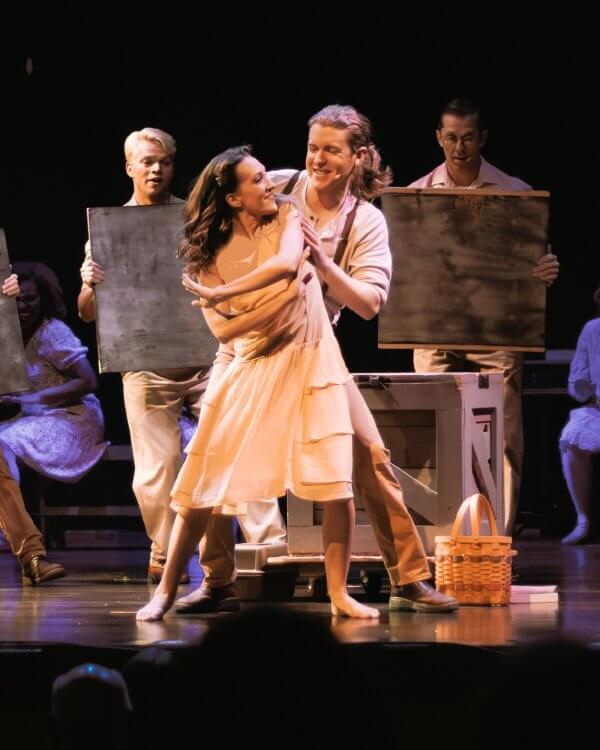 Actors perform a lively dance scene on stage, with two in the foreground embracing, while others hold rectangular props in the background—a vibrant moment from Live Theater Memphis musicals and plays.