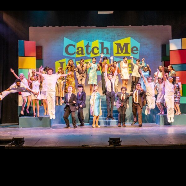A large group of performers in colorful costumes pose and jump on stage in front of a sign reading "Catch Me" during a live theater production, showcasing the excitement of musicals and plays in Memphis.