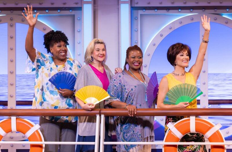 Four women stand on a ship deck holding colorful fans and waving, with life rings and a blue ocean in the background, reminiscent of the vibrant energy found in musicals and live theater performances.