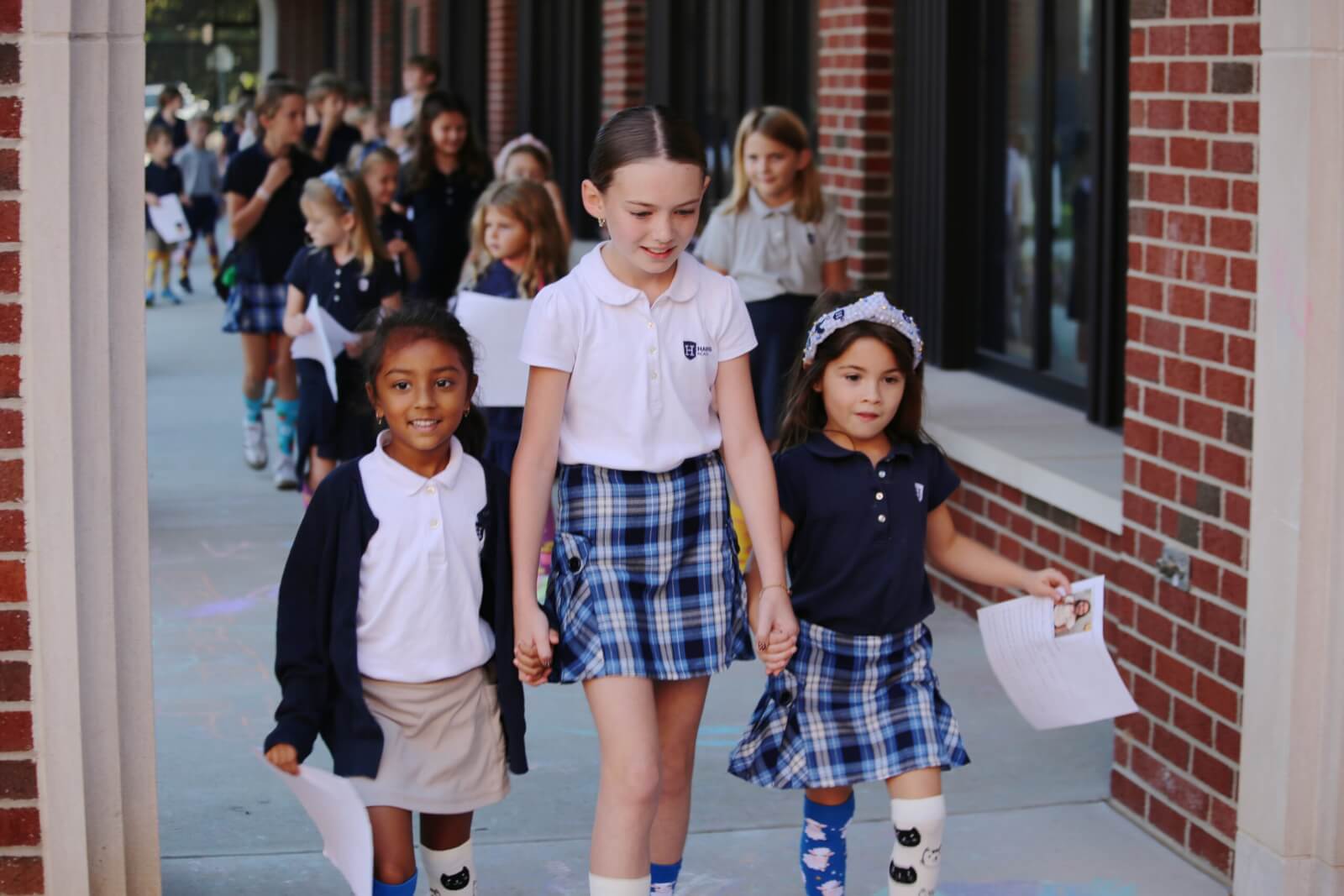 Nashville Private School Open House Dates - 6 A group of school children in uniforms from a Nashville private school walk outside along a brick building, with three girls holding hands at the front and others following behind.
