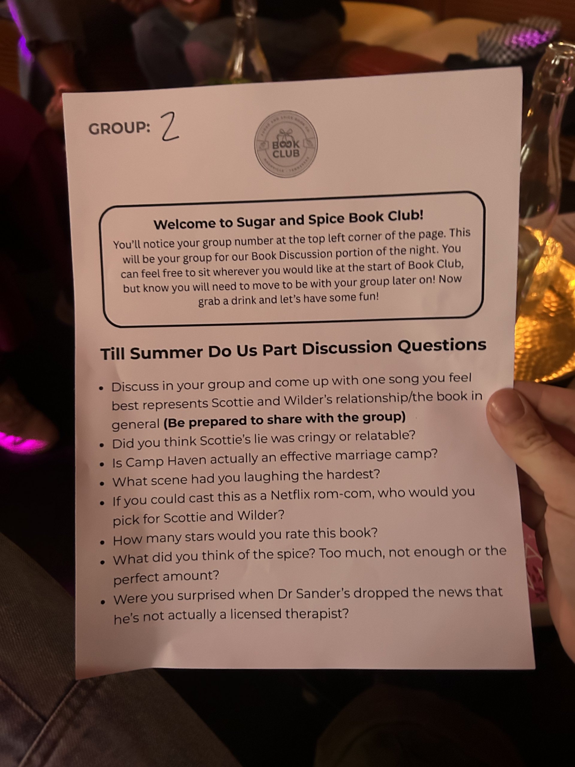 A hand holds a paper titled “Till Summer Do Us Part Discussion Questions” for Group 2 at a Book Club Nashville meeting, with Sugar & Spice Co drinks and dim lighting visible in the background.