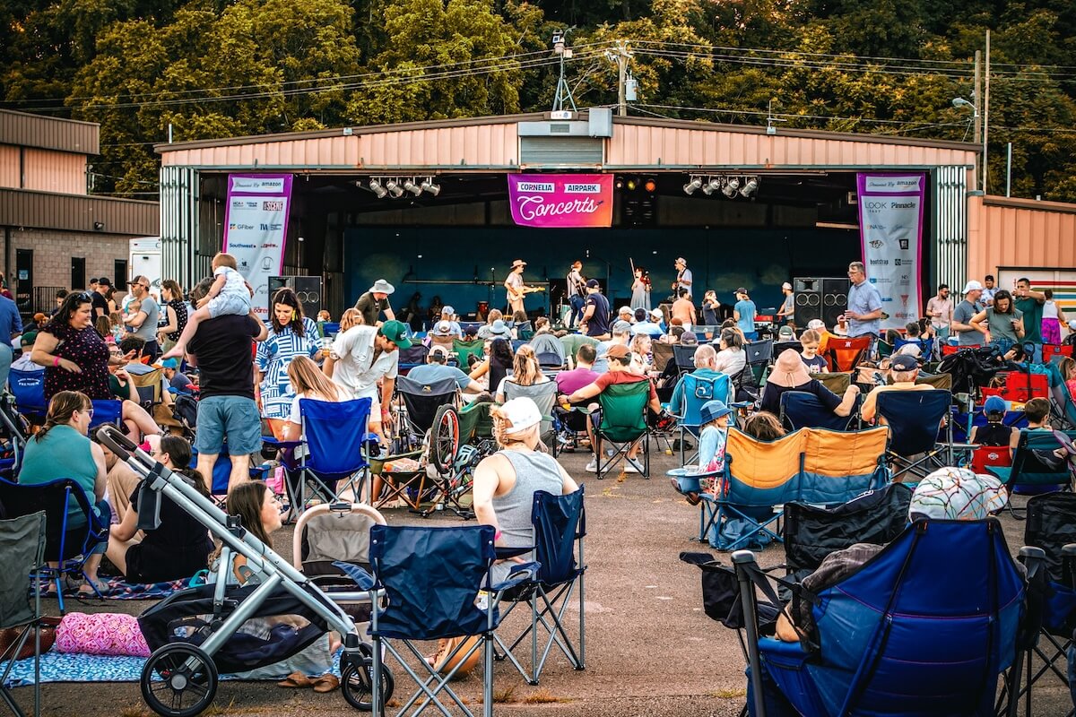 A crowd sits on folding chairs and blankets, enjoying a band perform on an outdoor stage as part of the Nashville Concert Series at a lively community concert event.