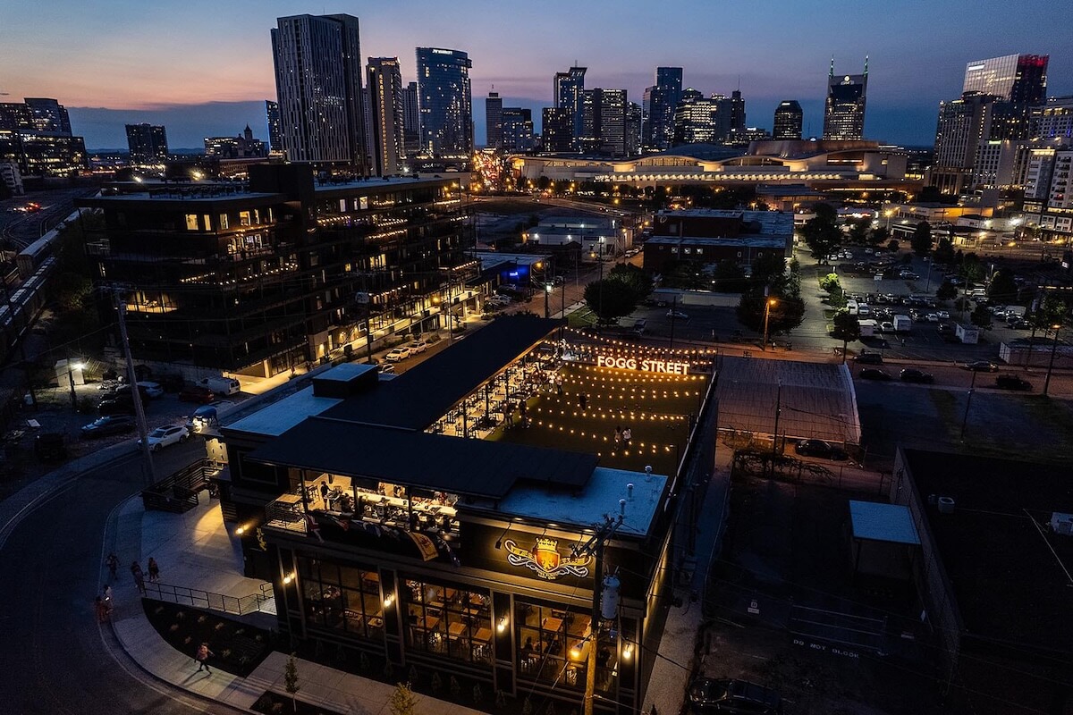 Aerial view of a rooftop restaurant with string lights, surrounded by city buildings at dusk, with a downtown skyline in the background—an ideal setting for enjoying a Nashville concert series under the stars.