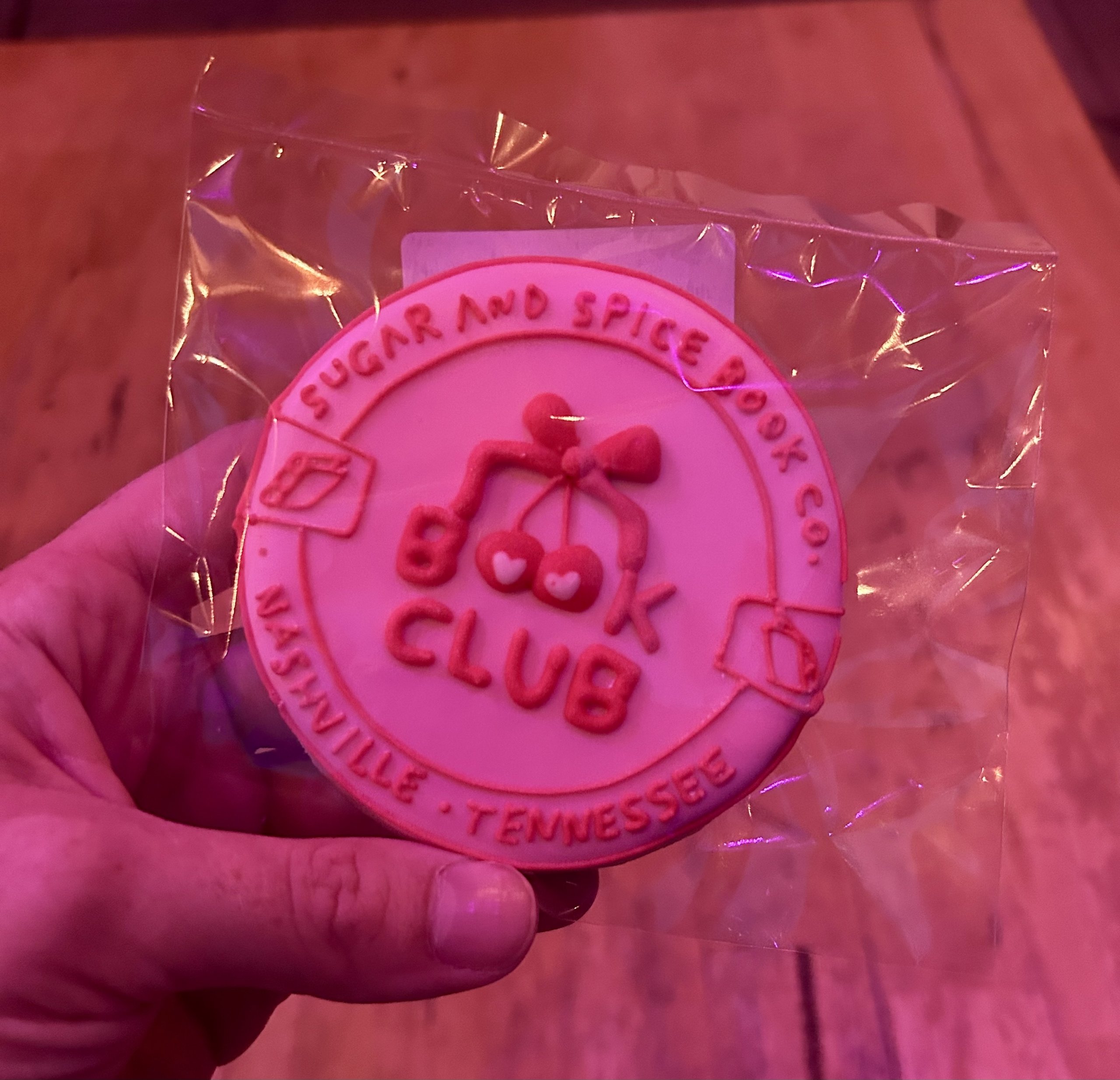 A hand holds a packaged cookie decorated with "Book Club," cherries, books, and the text "Sugar & Spice Co. Nashville, Tennessee"—the perfect treat for your next book club Nashville gathering.