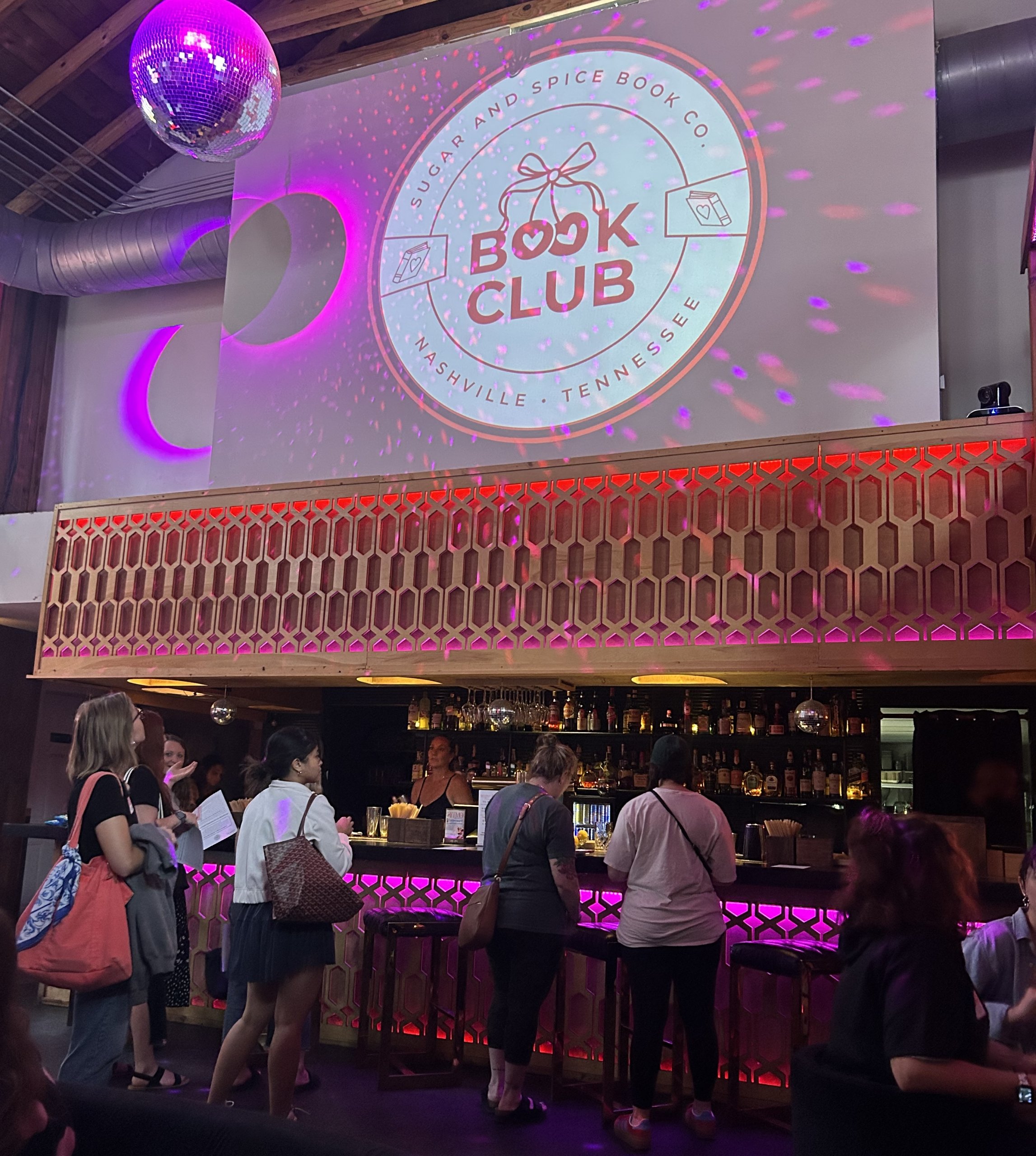 People stand in line at Sugar & Spice Co, a popular Book Club Nashville spot, with a large "Book Club" logo projected on the wall and a disco ball hanging from the ceiling.