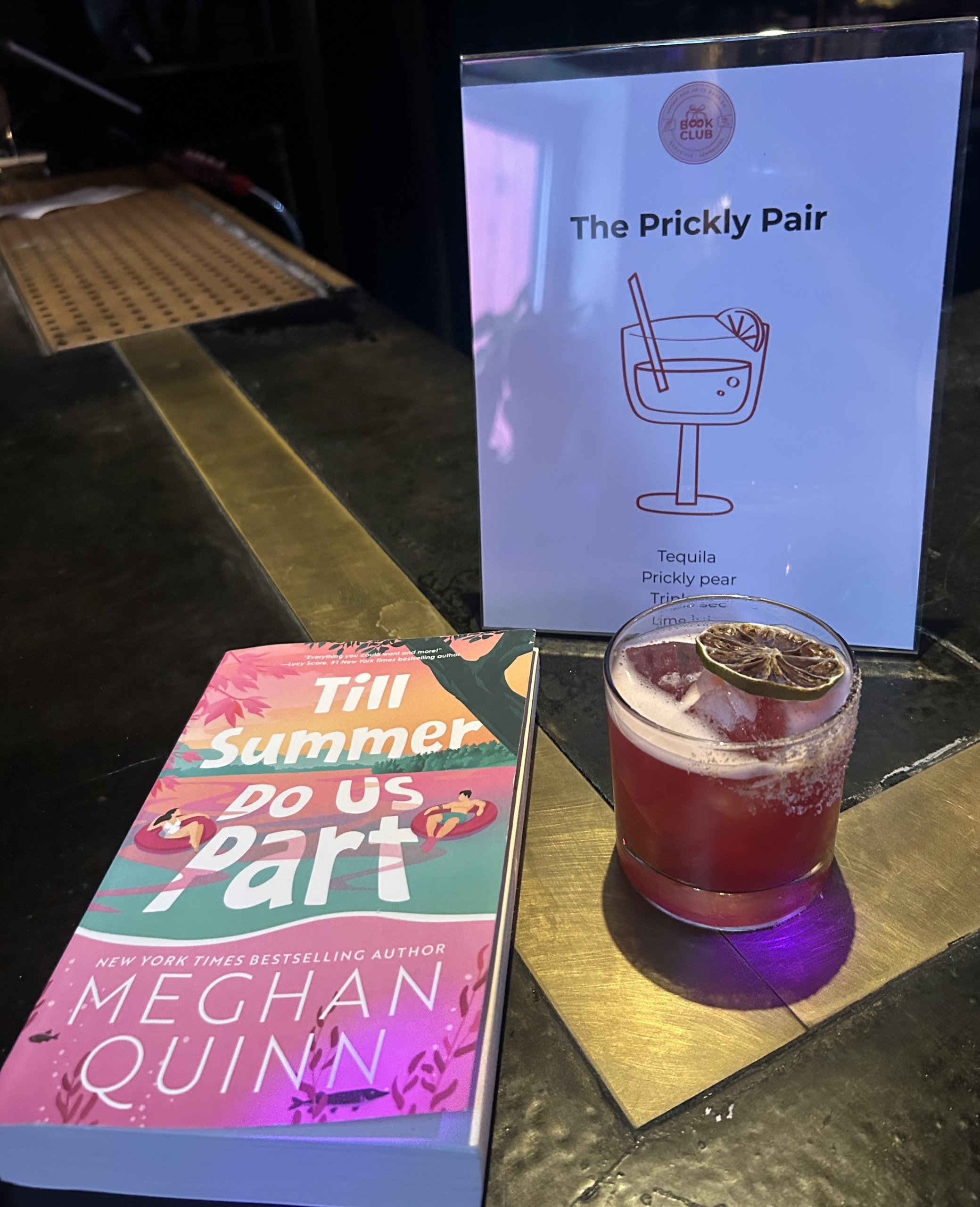 A pink cocktail from Sugar & Spice Co, garnished with a dried lime, sits beside the book "Till Summer Do Us Part" at Book Club Nashville, with a sign listing the drink "The Prickly Pair" and its ingredients.