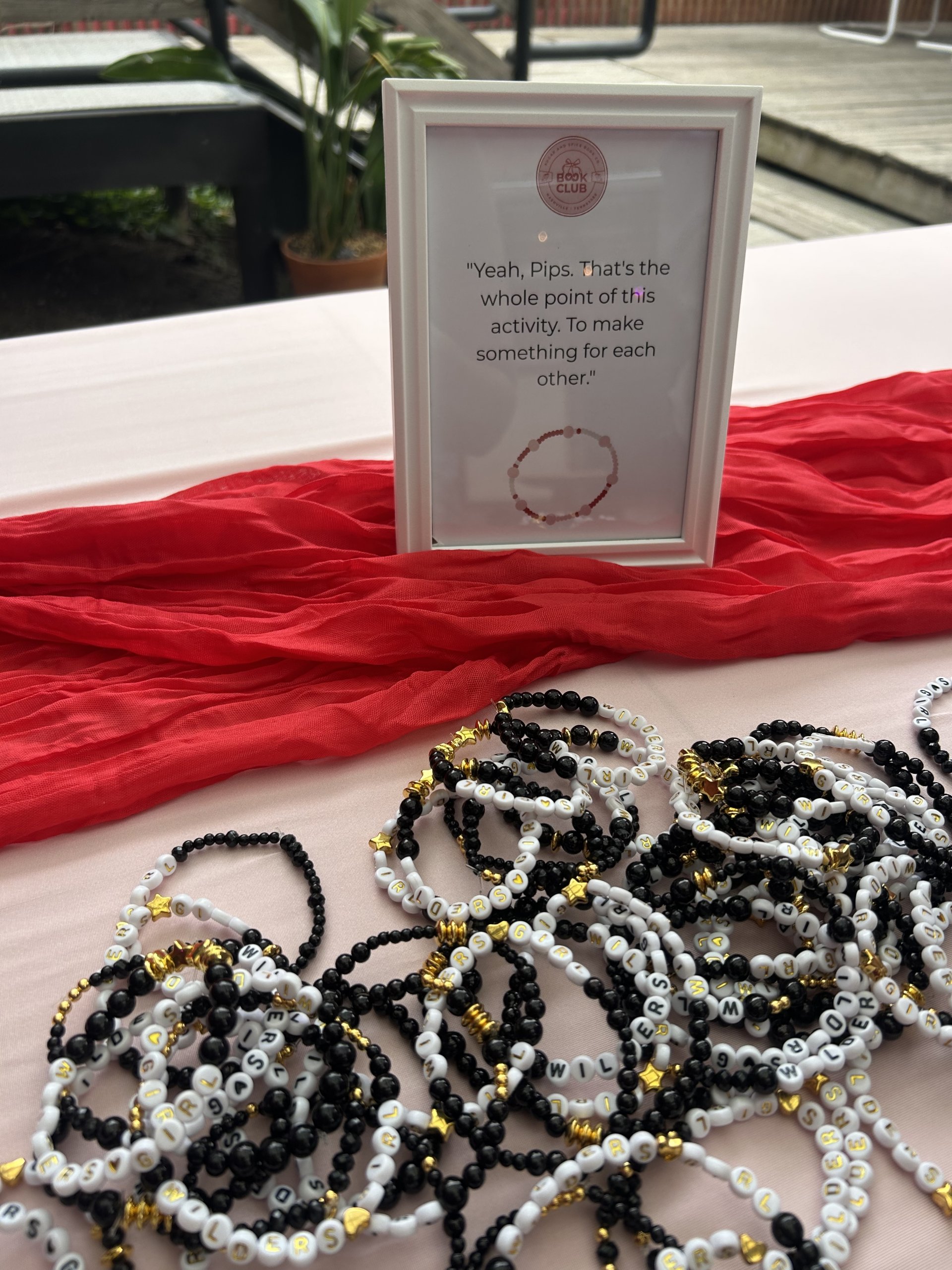 A table displays Sugar & Spice Co beaded bracelets in black, white, and gold next to a framed quote about making things for each other, with a red cloth and a potted plant in the background. Perfect for your book club Nashville gathering.