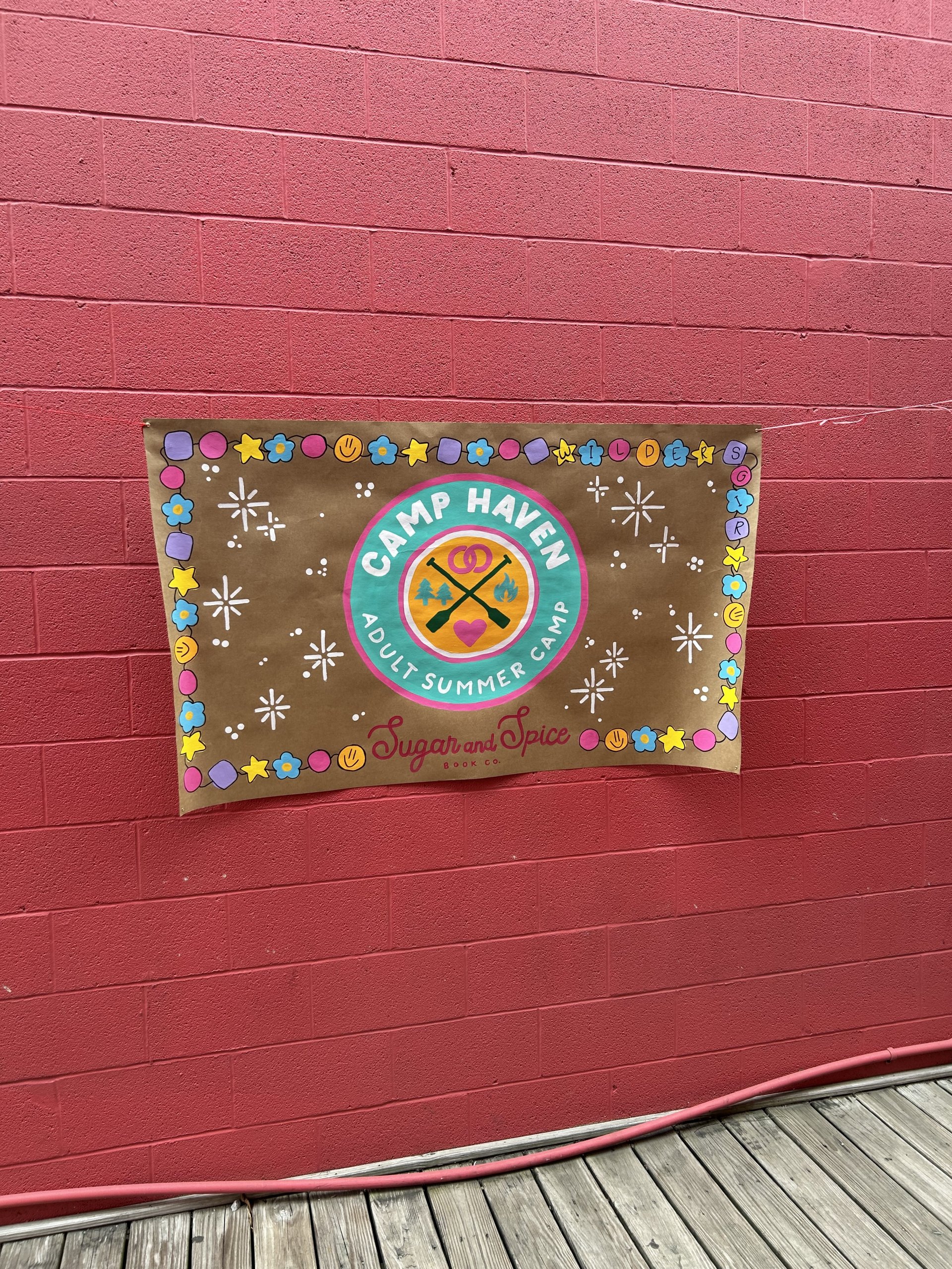 A colorful banner on a red brick wall reads "Camp Haven Adult Summer Camp @ Sugar & Spice Co.," decorated with stars, circles, and smiley faces—a vibrant scene perfect for Book Club Nashville gatherings.