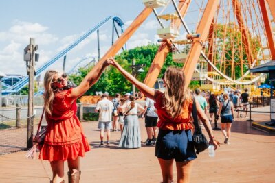 Two women holding hands and raising their arms while walking in an amusement park during one of the lively Southern events, with a large Ferris wheel and roller coaster visible in the background.