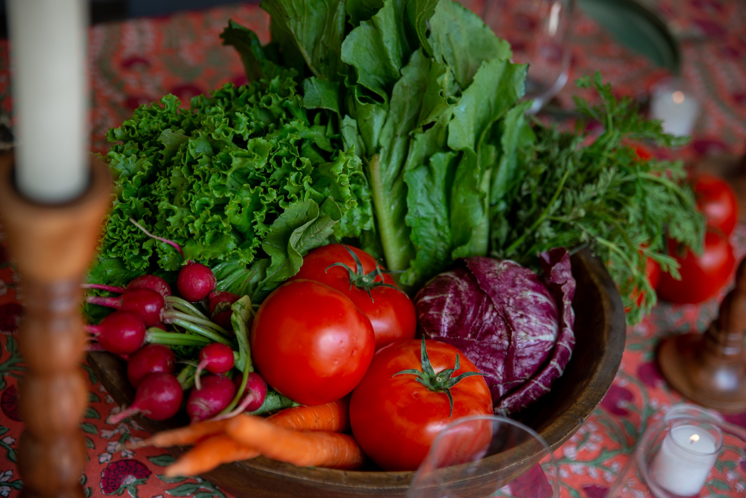 Veggies like radishes, lettuce, cabbages, tomatoes, and carrots in a wooden bowl on a beautiful floral tablecloth