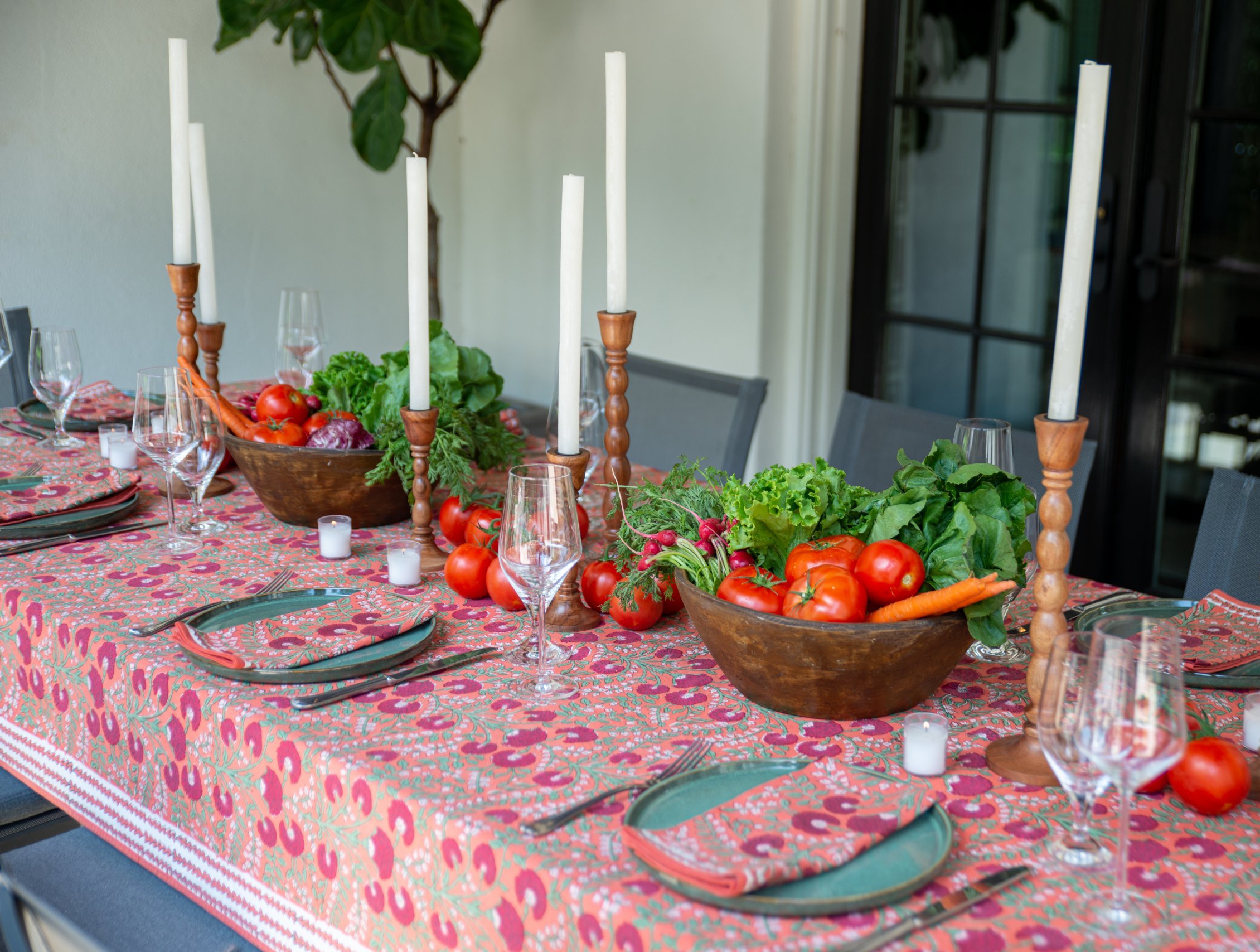 A pretty, summer tablescape featuring a salmon-colored floral tabletop with a wood bowl, wooden candlesticks, and bright vegetables