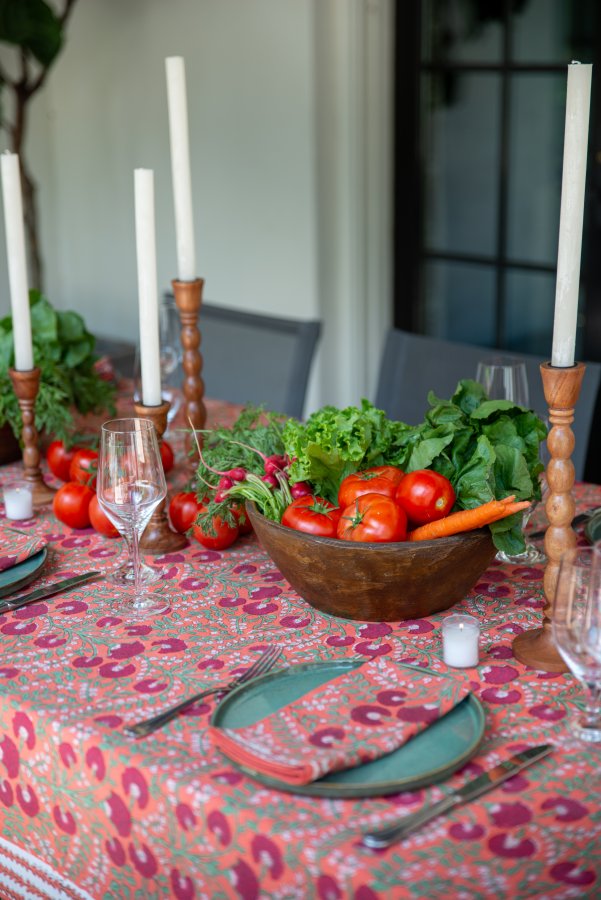 A pretty, summer tablescape featuring a salmon-colored floral tabletop with a wood bowl, wooden candlesticks, and bright vegetables