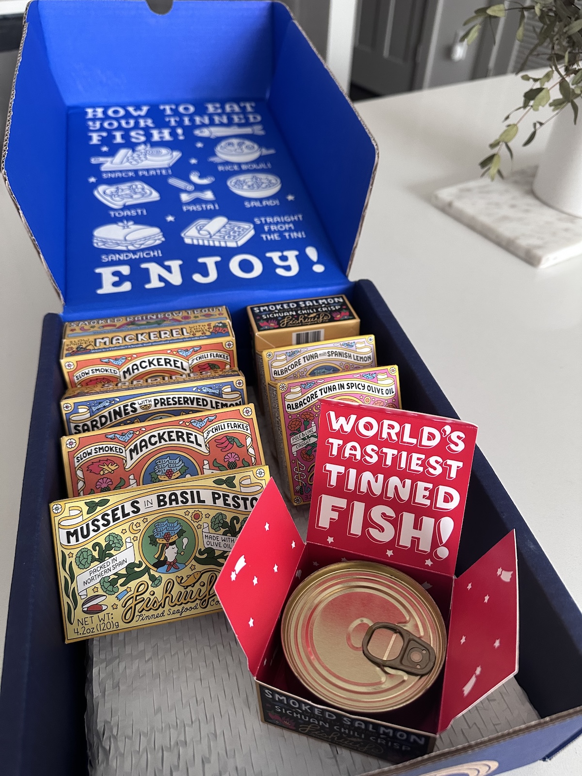 A selection of colorful tinned fish packages displayed in an open box with a sign reading "WORLD'S TASTIEST TINNED FISH!" and instructions for eating tinned fish—featured on the SB Hot List August.