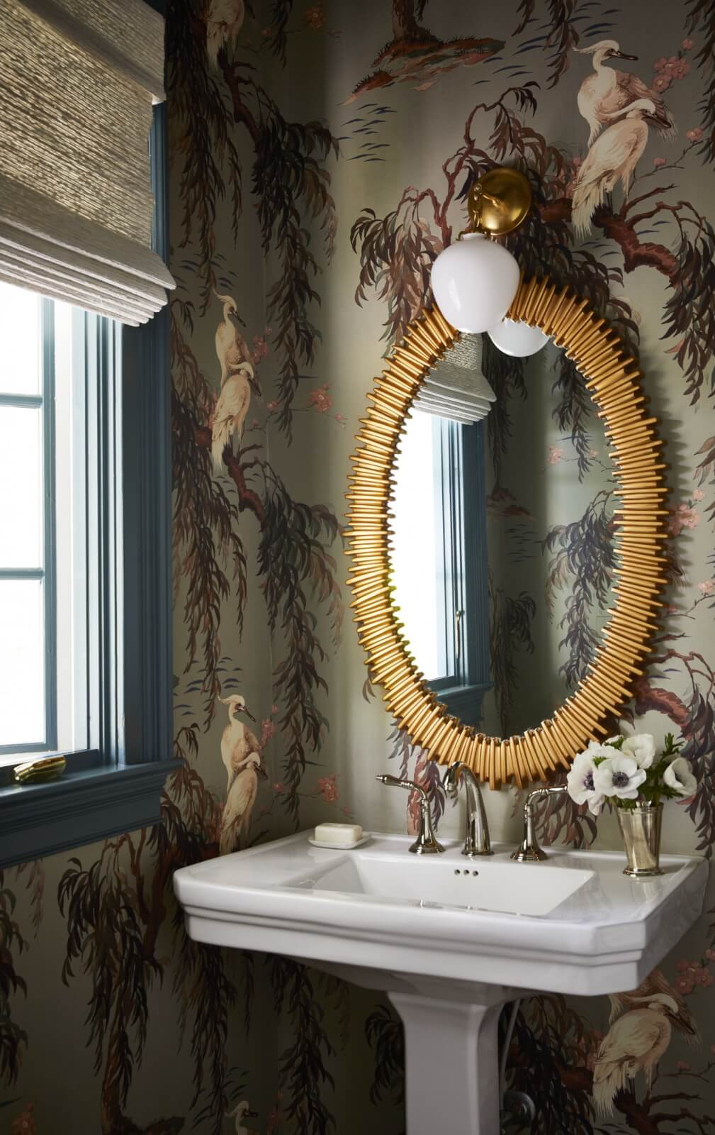Bathroom with a white pedestal sink, oval gold-framed mirror, floral wallpaper featuring birds, a window with a woven shade, and a vase of white flowers—reflecting classic Georgian architecture near Lake Wylie.
