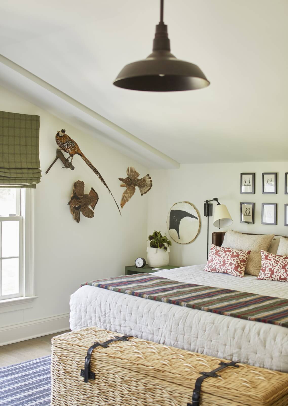 A bedroom with a bed, patterned bedding, wall-mounted bird taxidermy, framed art, a window with a green shade overlooking Lake Wylie, a wicker trunk, and a black pendant light.