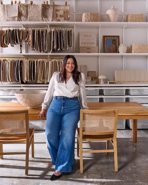 A woman in a white blouse and blue jeans stands, smiling, in Silo Studio—a modern design space with fabric samples, woven baskets, and wooden furniture displayed on shelves behind her.