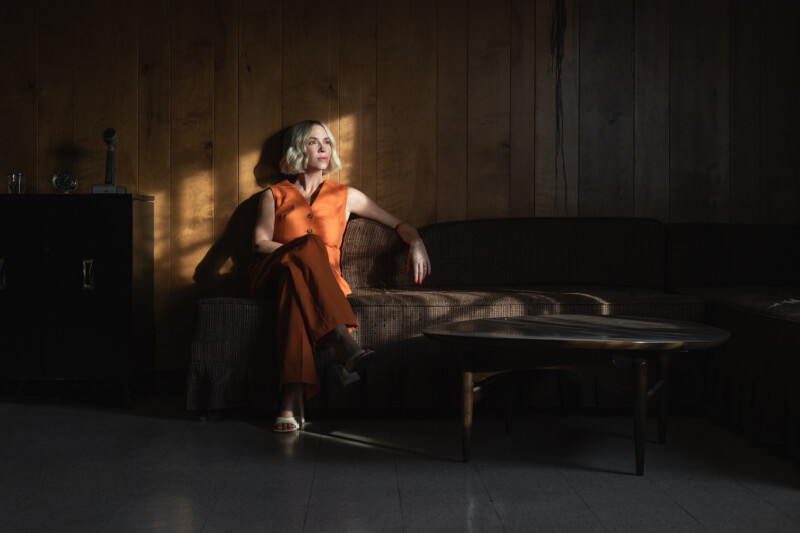A woman in an orange outfit sits on a dark couch in a wood-paneled room, with light streaming through a window and casting shadows on the wall.