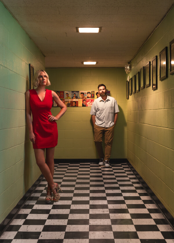 A woman in a red dress stands with one hand on her hip in a checkered hallway, while a man in a white shirt and tan pants leans against the back wall.