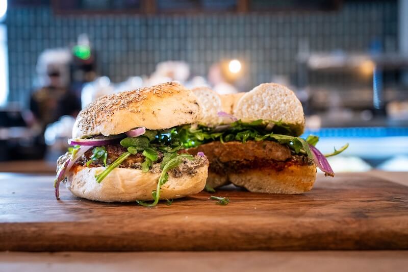 A sliced sandwich with a vegetable patty, arugula, red onions, and sauce on a seeded bun, displayed on a wooden board.