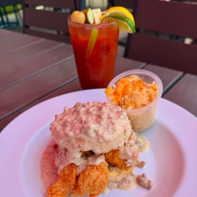 A plate with fried chicken topped with sausage gravy on a biscuit, mashed potatoes on the side, and a Bloody Mary garnished with lime, lemon, and pickles.
