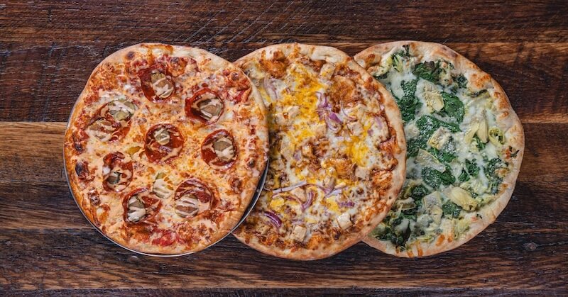 Three different pizzas on a wooden table: one with pepperoni, one with cheese and red onion, and one with spinach and artichoke.