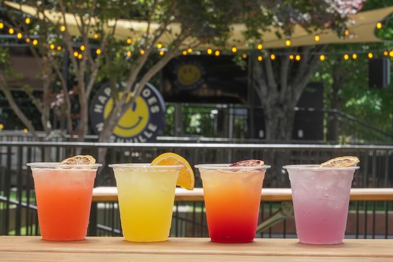 Four colorful cocktails in plastic cups, garnished with citrus slices, are lined up on a wooden table outdoors at one of the new Nashville restaurants, with trees and patio lights in the background.