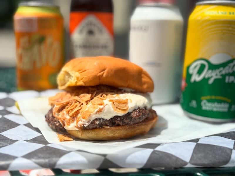 A cheeseburger topped with fried onions and sauce sits on checkered paper, with assorted canned drinks and bottles in the blurred background—a tasty pick for anyone exploring new Nashville restaurants in August 2025.