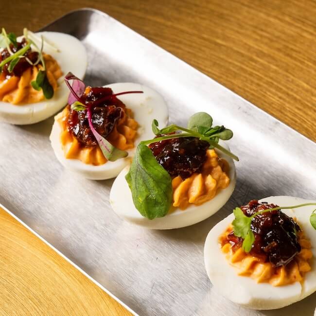 Four deviled eggs on a metal tray, topped with reddish spread, microgreens, and a dollop of dark chutney.