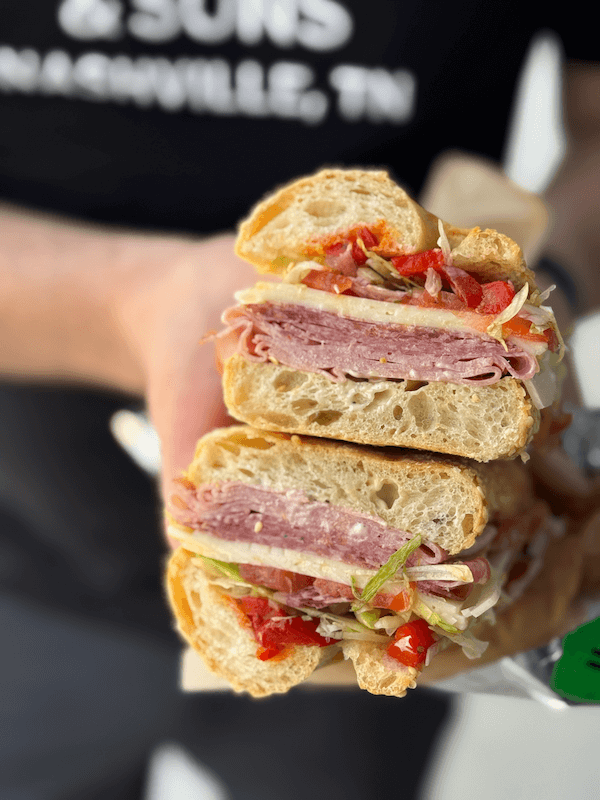 A person holds a sandwich cut in half, filled with deli meats, lettuce, tomatoes, and peppers on a crusty baguette.