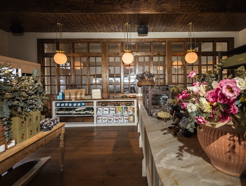 A bakery interior with wooden floors, display tables of products, a marble counter with flowers, and shelves stocked with baked goods and packaged items.