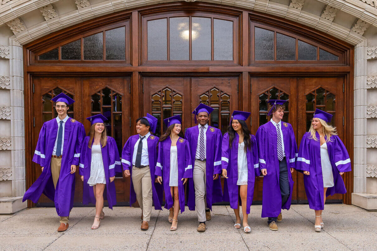 Seven graduates in purple caps and gowns stand and walk together outside a building with large wooden doors, smiling and celebrating after their achievements at a Nashville private school.