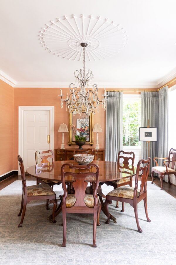 A formal dining room in this Buckhead home features a round wooden table, six chairs, chandelier, sideboard, lamps, framed art, and a large window with blue curtains—perfect for elegant Atlanta gatherings.