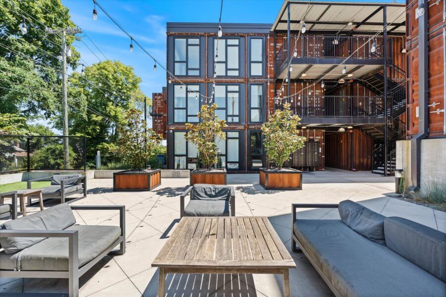 Outdoor patio area with modern seating and wooden tables, set in front of a multi-story building made from repurposed shipping containers at 2126 Byrum Ave, showcasing unique Nashville real estate on a sunny day.