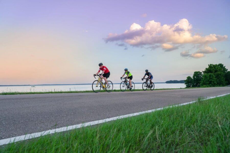 8 Mississippi Destinations Worth the Drive - 9 Three cyclists ride on a paved road near a body of water, with green grass and a colorful sky in the backgroundβan ideal scene for exploring top biking destinations in the South.