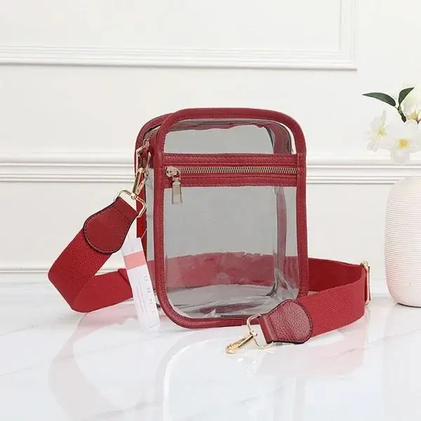 Clear rectangular bag with red trim, gold zipper, and adjustable red shoulder strap—one of our favorite Birmingham finds this September—placed on a white surface next to a white vase with flowers.
