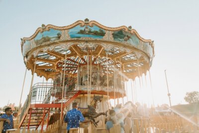 A double-decker carousel with people riding and standing nearby, surrounded by a wooden fence, under a clear sky with bright sunlight—a charming scene reminiscent of Birmingham events in October.