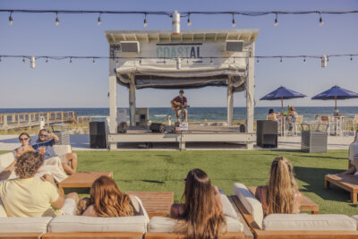 A musician performs on an outdoor stage by the ocean, while people sit on lounge chairs and watch under string lights—creating a scene that captures the essence of the best fall vacations.